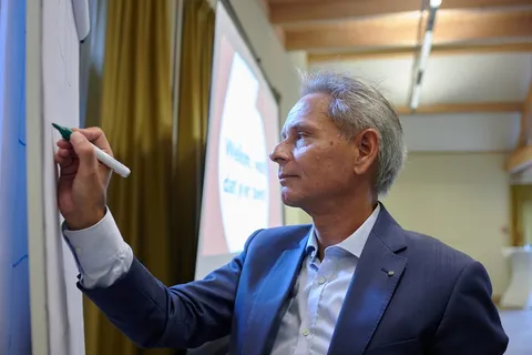 Man schrijft met een stift op een flipover, op de achtergrond een scherm met de tekst 'Welkom, wat fijn dat je er bent'