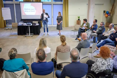 Twee sprekers openen de bijeenkomst voor het publiek, met op het scherm 'Welkom, wat fijn dat je er bent!'