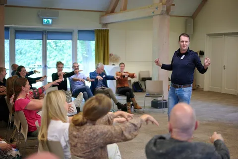 Workshopleider doet een handgebaar voor, deelnemers in een kring doen mee