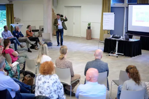 Spreker licht een dia met de tekst 'De aanpak' toe voor een zittend publiek
