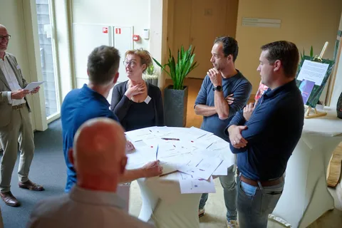 Groep deelnemers bespreekt papieren staand rond een sta-tafel