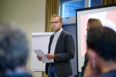Man met een blaadje papier spreekt de zaal toe voor een scherm