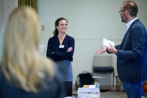 Vrouw met gekruiste armen glimlacht terwijl een collega iets uitlegt