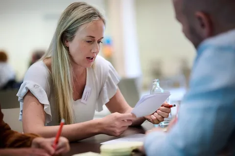 Vrouw legt een collega iets uit met papier in de hand aan tafel