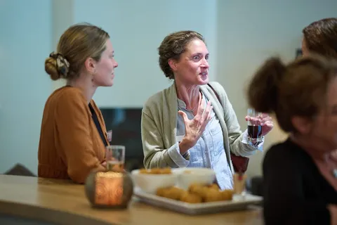 Vrouw praat gebarend met een drankje tijdens de borrel bij een schaal bittergarnituur