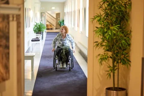 Vrouw in rolstoel rijdt door een gang met planten langs de kamers